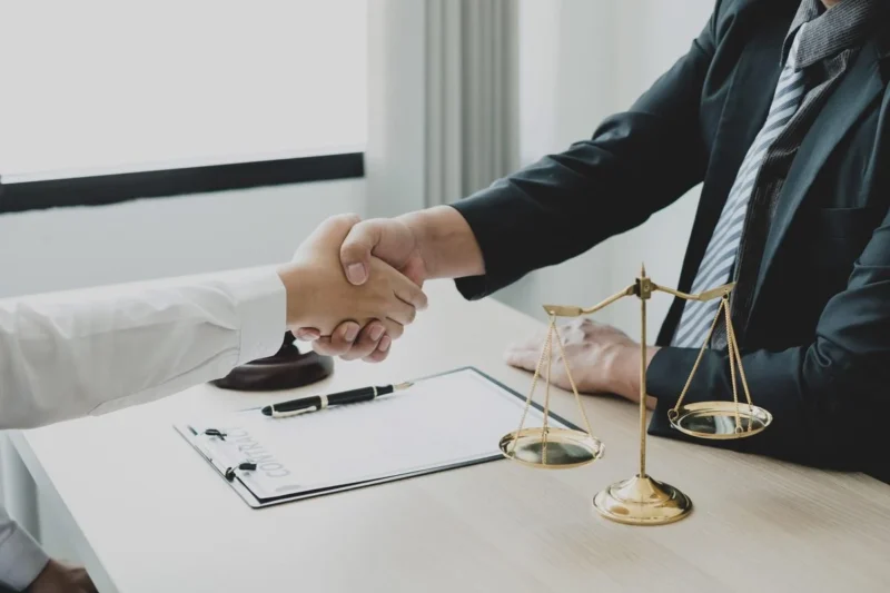 A handshake across a desk beside brass scales of justice and a gavel
